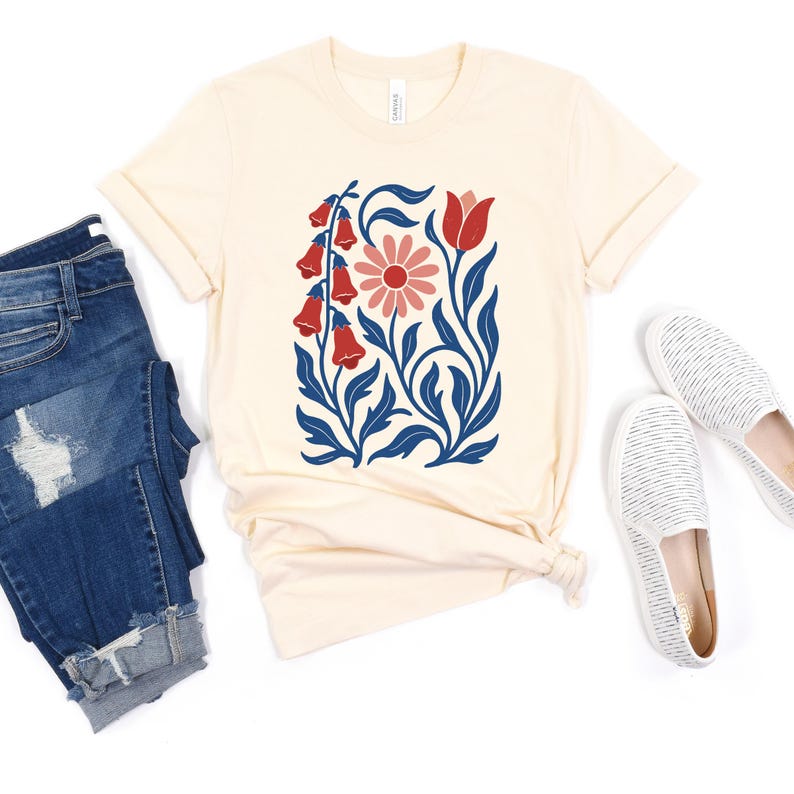 May include: Cream-colored t-shirt featuring a floral design with red bell-shaped flowers, a pink daisy, and blue leaves. The shirt is paired with blue jeans and white striped slip-on shoes, creating a casual, everyday look.