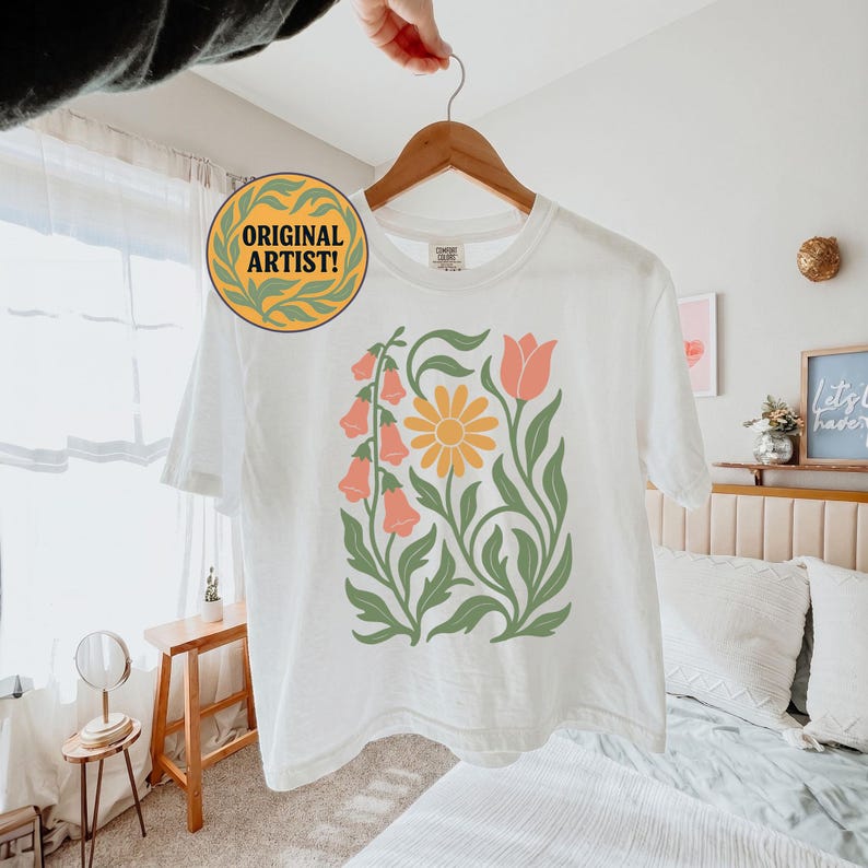 May include: A white short-sleeve t-shirt featuring a central graphic print of a yellow daisy, pink bell-shaped flowers, and a single pink tulip, all connected by green stems and leaves. A circular badge with the text ORIGINAL ARTIST is attached.