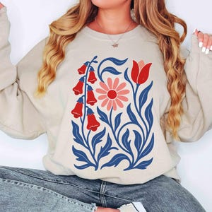 May include: Beige sweatshirt featuring a floral design. The graphic includes red bell-shaped flowers, a pink daisy, and a red tulip, all set against blue leaves and stems. The sweatshirt is a neutral color, suitable for casual wear.