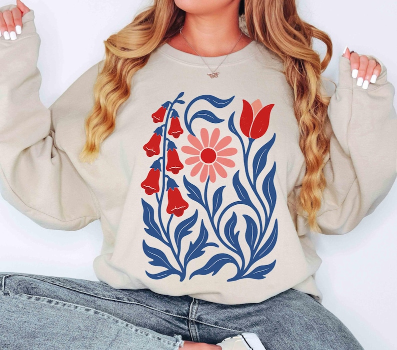May include: Beige sweatshirt featuring a floral design. The graphic includes red bell-shaped flowers, a pink daisy, and a red tulip, all set against blue leaves and stems. The sweatshirt is a neutral color, suitable for casual wear.