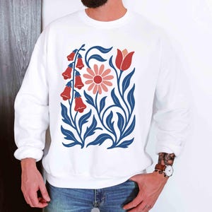 May include: White sweatshirt featuring a floral design with red bell-shaped flowers, a pink daisy, and blue leaves and stems. The design is centered on the front of the long-sleeved top. The sweatshirt is made of a soft material.