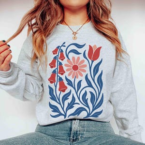 May include: A light gray sweatshirt featuring a floral design. The design includes red bell-shaped flowers, a pink daisy, and blue leaves and stems. The sweatshirt is a casual, comfortable style.