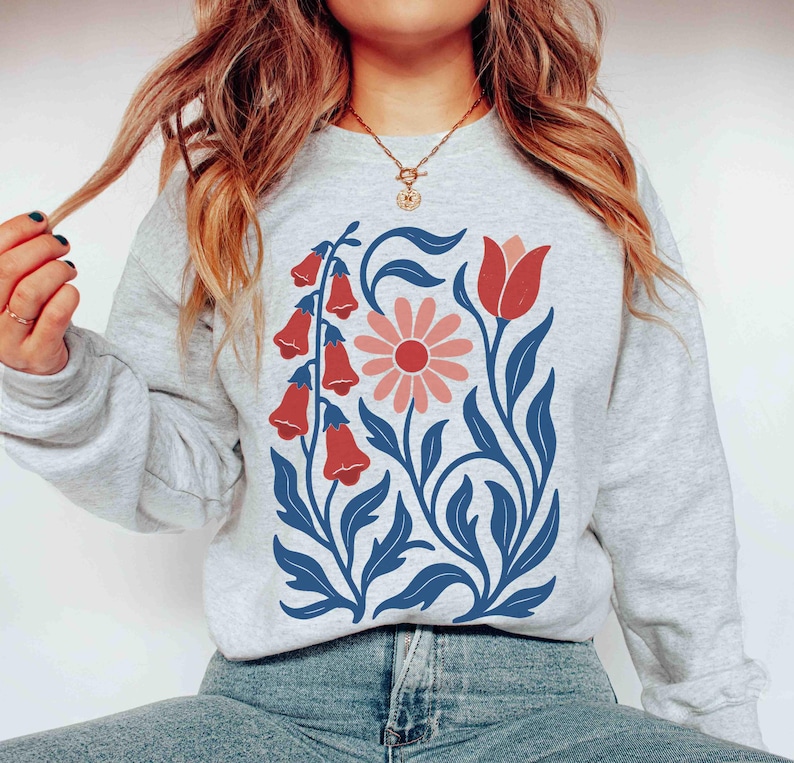 May include: A light gray sweatshirt featuring a floral design. The design includes red bell-shaped flowers, a pink daisy, and blue leaves and stems. The sweatshirt is a casual, comfortable style.