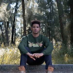May include: A dark green sweatshirt with a mushroom and floral graphic print. The sweatshirt has a crew neck and long sleeves.