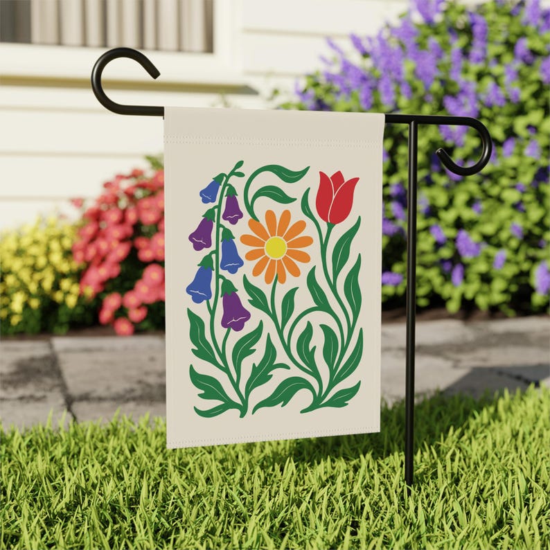 May include: A cream-colored garden flag featuring a floral design with a daisy, tulip, and bell-shaped flowers in orange, red, blue, and purple. The flag is displayed on a black metal stand in a grassy yard, with colorful flowers in the background.