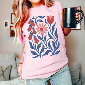 May include: A light pink t-shirt featuring a floral design. The design includes red bell-shaped flowers, a pink daisy, and blue leaves and stems. The shirt is a soft, comfortable material.