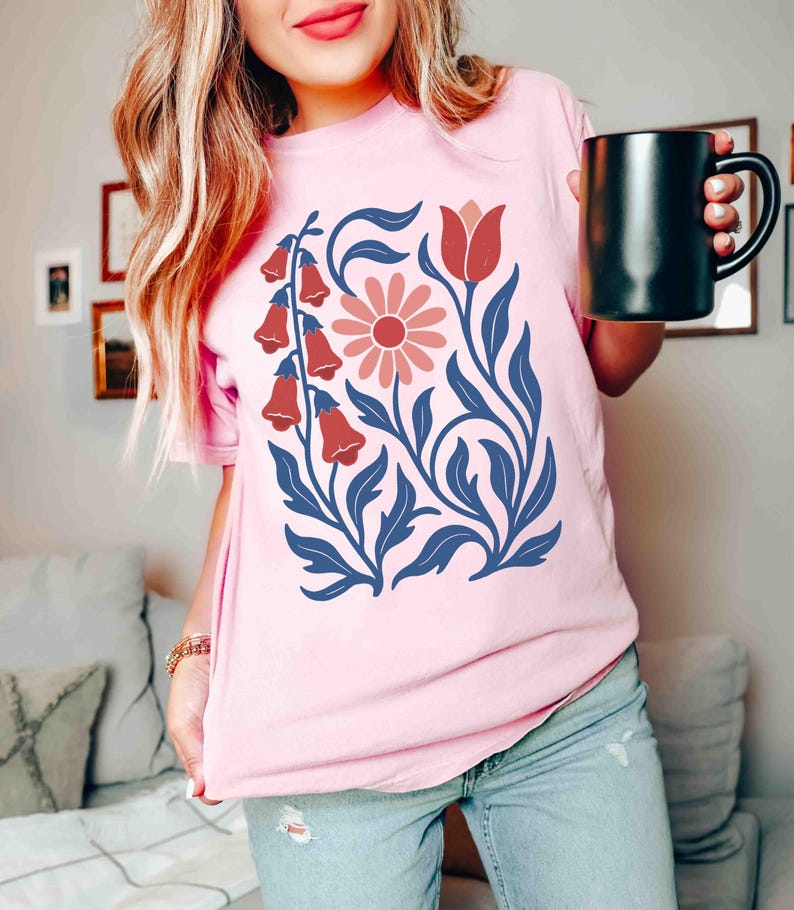 May include: A light pink t-shirt featuring a floral design. The design includes red bell-shaped flowers, a pink daisy, and blue leaves and stems. The shirt is a soft, comfortable material.