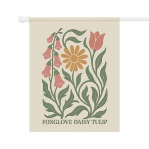 May include: A beige garden flag featuring a floral design with foxgloves, a daisy, and a tulip in shades of peach, orange, and green. The words "FOXGLOVE DAISY TULIP" are printed at the bottom in a dark green font. The flag is mounted on a white pole.