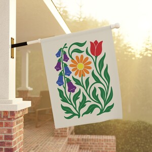 May include: A cream-colored garden flag featuring a floral design. The design includes a red tulip, an orange daisy, and purple and blue bell-shaped flowers, all with green stems and leaves. The flag is hanging outside on a white pole.
