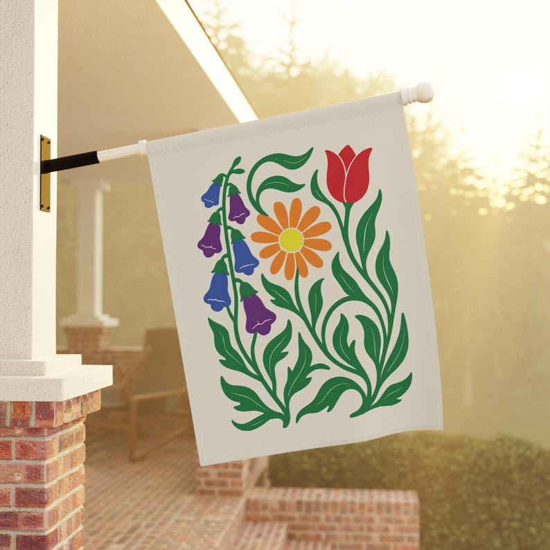 May include: A cream-colored garden flag featuring a floral design. The design includes a red tulip, an orange daisy, and purple and blue bell-shaped flowers, all with green stems and leaves. The flag is hanging outside on a white pole.