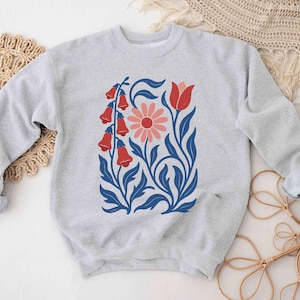May include: A light gray sweatshirt featuring a floral design. The design includes red bell-shaped flowers, a pink daisy, and blue leaves and stems. The sweatshirt has long sleeves with rolled-up cuffs.