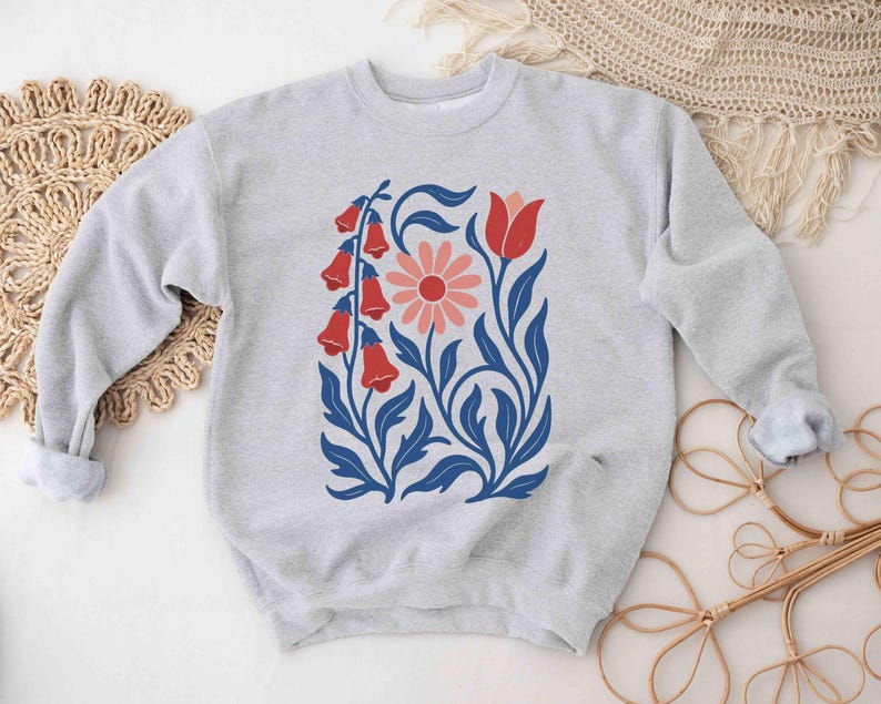 May include: A light gray sweatshirt featuring a floral design. The design includes red bell-shaped flowers, a pink daisy, and blue leaves and stems. The sweatshirt has long sleeves with rolled-up cuffs.
