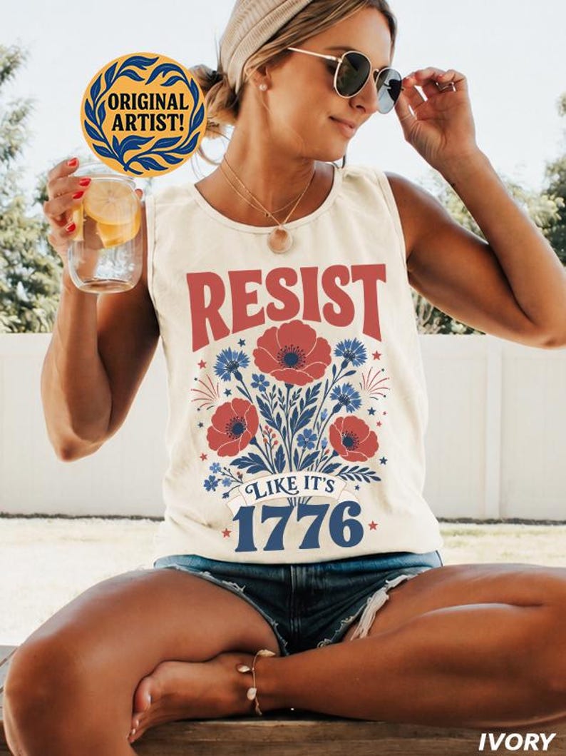 May include: An ivory sleeveless tank top featuring a graphic print with red poppies and blue cornflowers. The text reads RESIST in red, and LIKE IT'S 1776 in blue. The design includes small blue stars and red firework-like accents on the front.