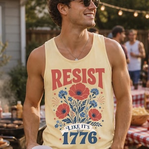 May include: A pale yellow tank top featuring a graphic print with red poppies and blue cornflowers. The text reads RESIST in red letters above the flowers, and LIKE IT'S 1776 in blue letters below the floral design, accented with small red stars.