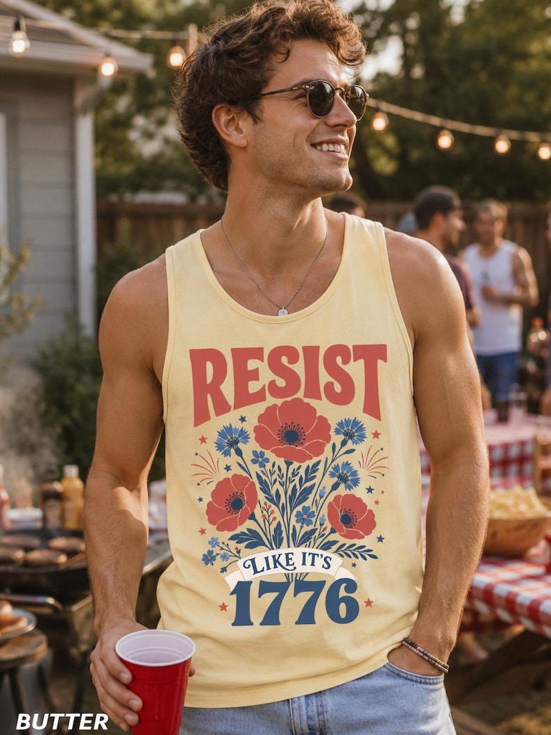 May include: A pale yellow tank top featuring a graphic print with red poppies and blue cornflowers. The text reads RESIST in red letters above the flowers, and LIKE IT'S 1776 in blue letters below the floral design, accented with small red stars.