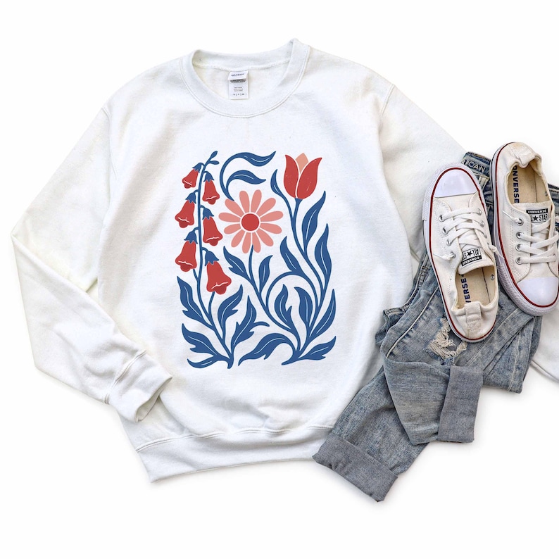May include: White sweatshirt featuring a floral design with red, pink, and blue elements. The design includes a large pink flower, red bell-shaped flowers, and blue leaves and stems. The sweatshirt is paired with blue jeans and white canvas sneakers.