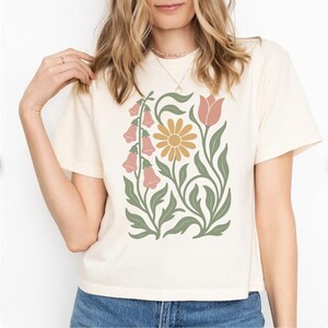 May include: A cream-colored short-sleeve t-shirt featuring a central graphic print of stylized botanical elements, including pink bell-shaped flowers, a yellow daisy-like flower, and a single pink tulip, all accented with sage green leaves.