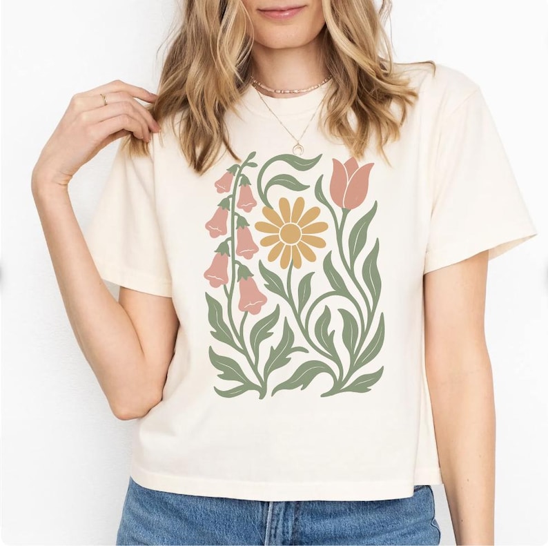 May include: A cream-colored short-sleeve t-shirt featuring a central graphic print of stylized botanical elements, including pink bell-shaped flowers, a yellow daisy-like flower, and a single pink tulip, all accented with sage green leaves.