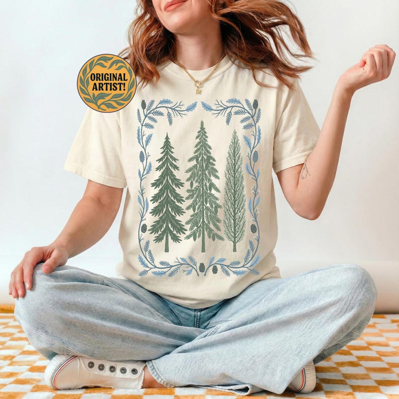 May include: Cream-colored t-shirt featuring a nature-themed design. The graphic includes three green pine trees framed by a blue and green floral border. The text "ORIGINAL ARTIST!" is in a gold circle.