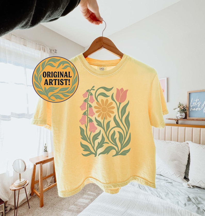 May include: A pale yellow short-sleeve t-shirt featuring a central graphic of a large yellow daisy, pink bell-shaped flowers, and a single pink tulip, all surrounded by green leafy stems. A circular badge with the text ORIGINAL ARTIST! is visible.