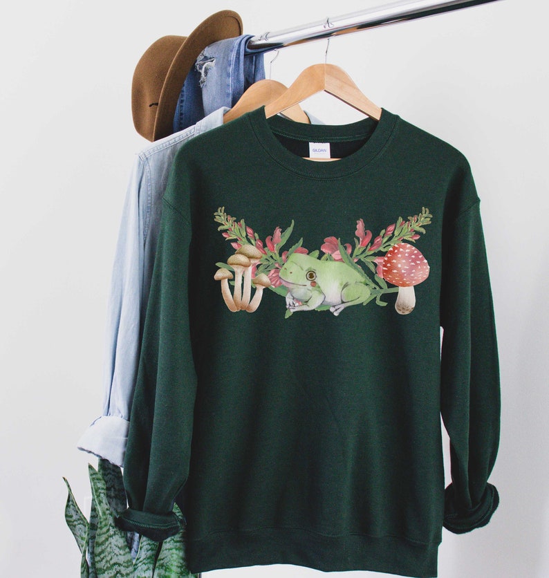 May include: A dark green sweatshirt with a white frog, red and white mushrooms, and pink flowers printed on the front.