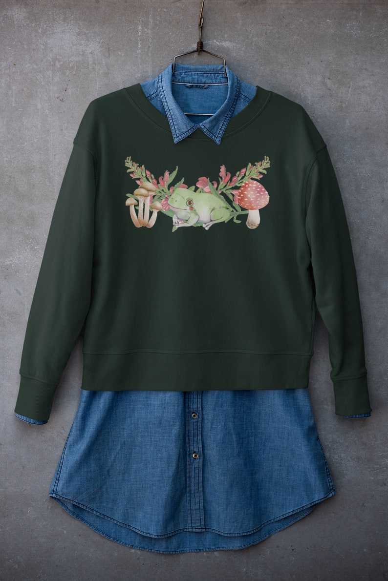 May include: A dark green sweatshirt with a watercolor design of a frog sitting on a branch with mushrooms and flowers.