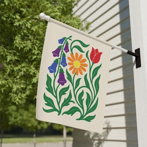 May include: A cream-colored garden flag featuring a floral design. The design includes a central orange daisy with a yellow center, a red tulip, and purple and blue bell-shaped flowers. Green leaves and stems complete the design.
