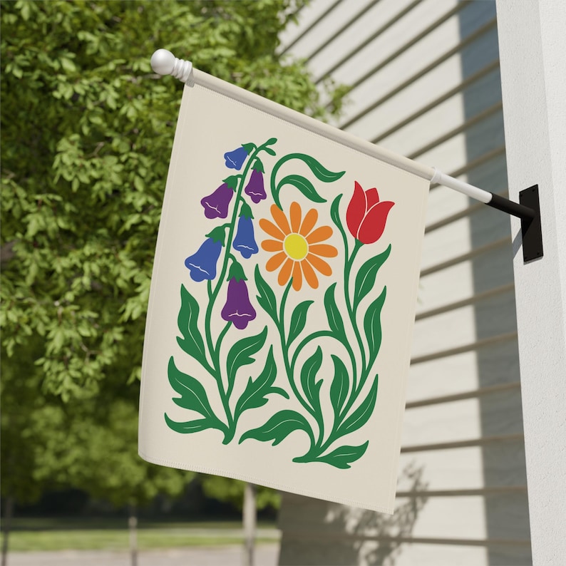 May include: A cream-colored garden flag featuring a floral design. The design includes a central orange daisy with a yellow center, a red tulip, and purple and blue bell-shaped flowers. Green leaves and stems complete the design.