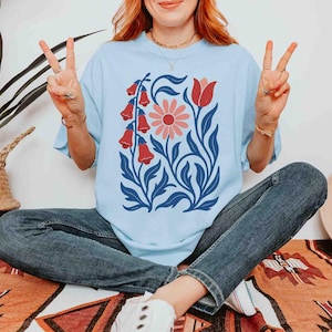 May include: Light blue t-shirt featuring a floral design with red bell-shaped flowers, a pink daisy, and a red tulip, set against blue leaves. The shirt is worn with blue jeans. A straw hat and a potted plant are in the background.