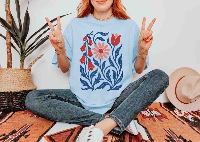 May include: Light blue t-shirt featuring a floral design with red bell-shaped flowers, a pink daisy, and a red tulip, set against blue leaves. The shirt is worn with blue jeans. A straw hat and a potted plant are in the background.