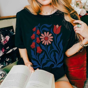 May include: Black t-shirt featuring a floral design with red bell-shaped flowers, a pink daisy, and blue leaves and stems. The design is centered on the shirt. The shirt is a short-sleeved crew neck.