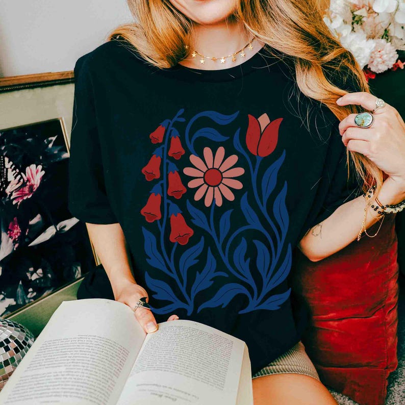 May include: Black t-shirt featuring a floral design with red bell-shaped flowers, a pink daisy, and blue leaves and stems. The design is centered on the shirt. The shirt is a short-sleeved crew neck.