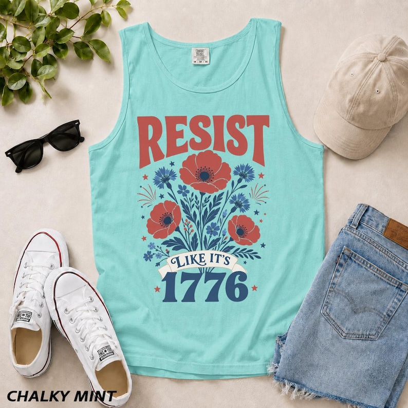 May include: A light turquoise tank top featuring a graphic print with the red text RESIST above a bouquet of red poppies and blue cornflowers, with the text LIKE IT'S 1776 in blue below the flowers, all centered on the front of the garment.