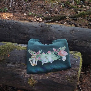 May include: A dark green sweatshirt with a frog, mushrooms, and flowers printed on the front. The sweatshirt is folded and lying on a log in a forest setting.