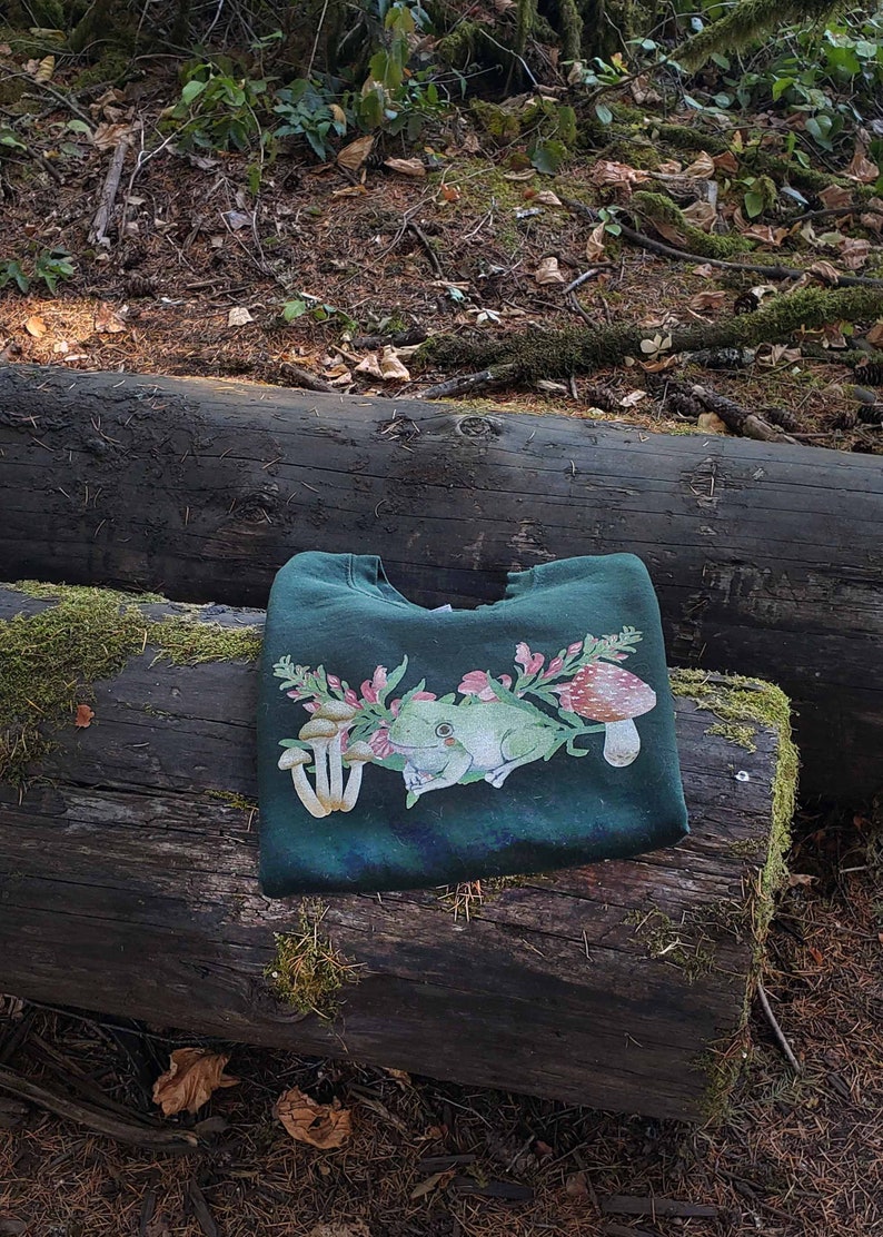 May include: A dark green sweatshirt with a frog, mushrooms, and flowers printed on the front. The sweatshirt is folded and lying on a log in a forest setting.