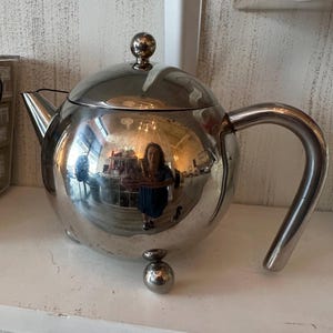 May include: A polished silver-tone metal teapot with a spherical body, a curved handle, a short spout, and a lid topped with a small spherical knob. The teapot stands on three small, round, ball-shaped feet and features a highly reflective surface.