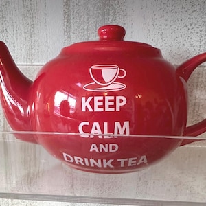 Peut inclure: Une théière en céramique rouge brillant avec un couvercle, ornée d'un motif blanc représentant une tasse sur une soucoupe au-dessus du texte blanc KEEP CALM AND DRINK TEA. La théière possède une anse courbée et un long bec verseur.