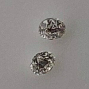 May include: Two small, round, clear, faceted gemstones with a silver setting. The gemstones are on a white surface.