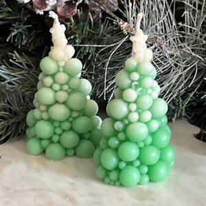 May include: Two green Christmas tree-shaped candles with a bubbly texture. The candles have a gradient of green hues, with a lighter shade at the top. The candles are placed on a white surface with pine needles and a pine cone in the background.