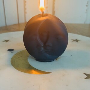 Puede incluir: Una vela esférica de color gris oscuro con un diseño de cara de luna creciente tallada. La vela está encendida, con una llama amarilla brillante. La vela se encuentra sobre un plato blanco con diseños de luna creciente y estrellas doradas.