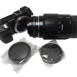 May include: A black Sony digital camera with a Sigma lens attached. The lens has measurement markings. Two lens caps are in plastic packaging. The camera and lens are on a white background.