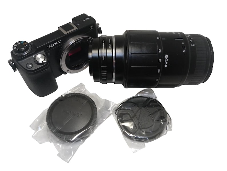 May include: A black Sony digital camera with a Sigma lens attached. The lens has measurement markings. Two lens caps are in plastic packaging. The camera and lens are on a white background.