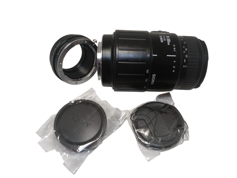 May include: A black Sigma 18-50mm f/3.5-5.6 DC lens with a lens adapter and two lens caps. The lens has measurement markings and the word "SIGMA" printed on it. The lens caps are black and one is labeled "SONY".