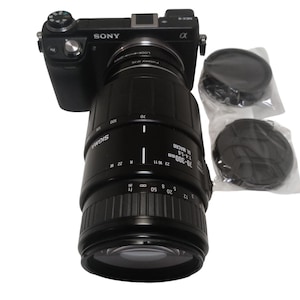 May include: A black Sony Alpha camera with a Sigma 70-300mm macro lens attached. Two lens caps are included. The lens has measurement markings. The camera and lens are set against a white background.