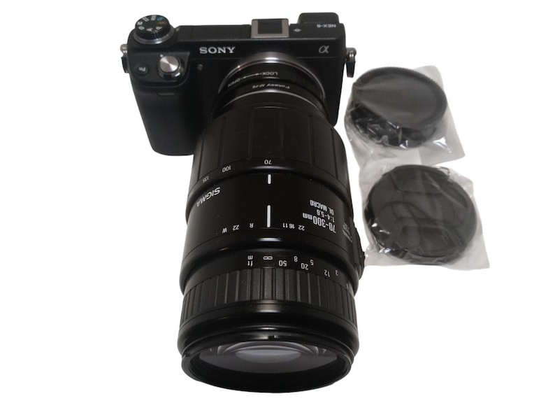 May include: A black Sony Alpha camera with a Sigma 70-300mm macro lens attached. Two lens caps are included. The lens has measurement markings. The camera and lens are set against a white background.