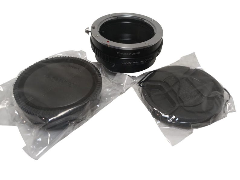 May include: A black and silver lens adapter with two black lens caps, all in clear plastic packaging. The adapter has the text "Fotasy M-R" and "LOCK" and "OPEN" labels. The caps are round and have a ridged edge.