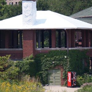May include: Exterior view of a brick building with a white roof and a tall chimney. The building has large windows and is surrounded by green foliage. A Coca-Cola vending machine is visible.