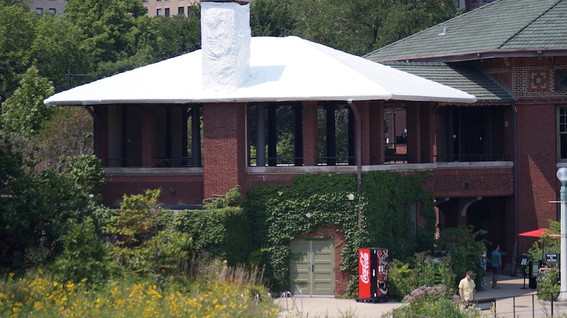 May include: Exterior view of a brick building with a white roof and a tall chimney. The building has large windows and is surrounded by green foliage. A Coca-Cola vending machine is visible.