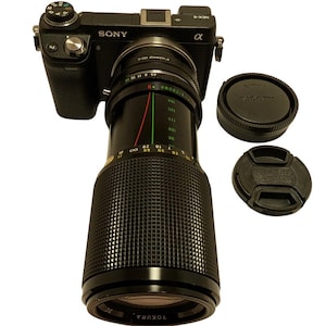 Puede incluir: Cámara Sony Alpha negra con un objetivo Tokura. El objetivo tiene un agarre texturizado y marcas de medición. También hay dos tapas de objetivo negras. La cámara está sobre un fondo blanco.
