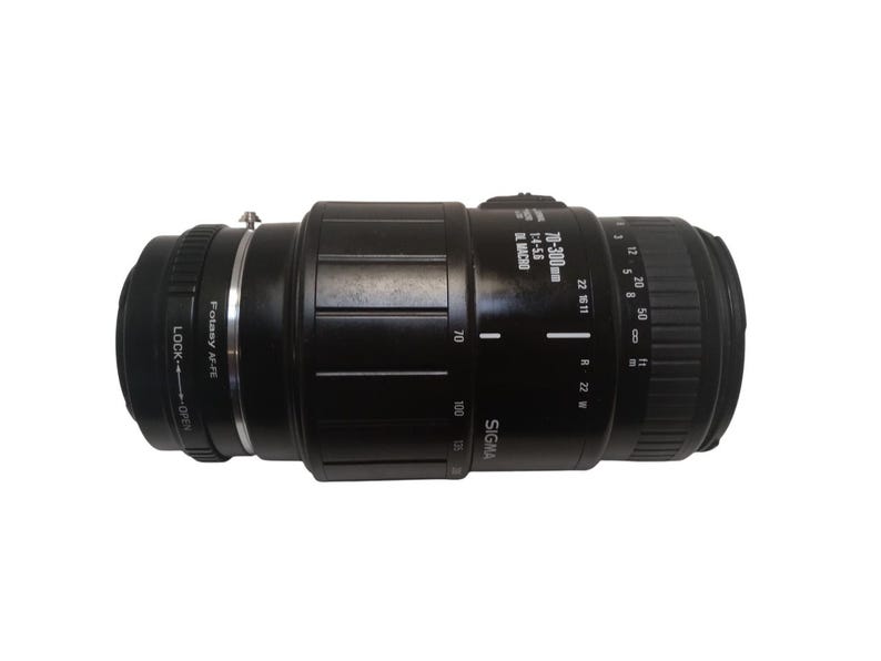 May include: A black Sigma 70-300mm f/4-5.6 macro zoom lens. The lens has markings for focal lengths, aperture settings, and a macro function. It features a Fotasy AF-FE mount and a lock/open mechanism.